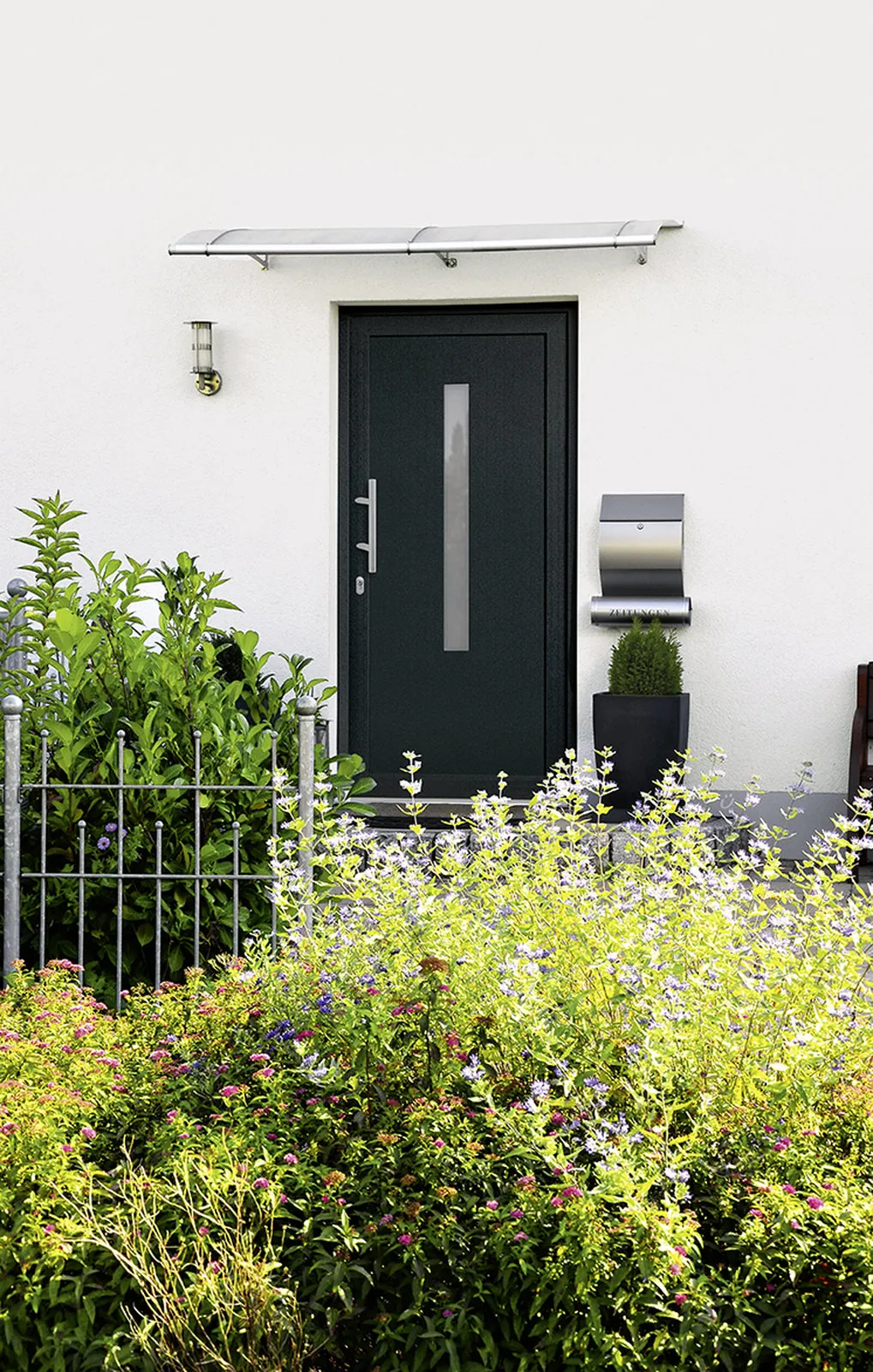 Anthrazitfarbene Haustür mit Glas-Vordach, Briefkasten und blühendem Vorgarten
