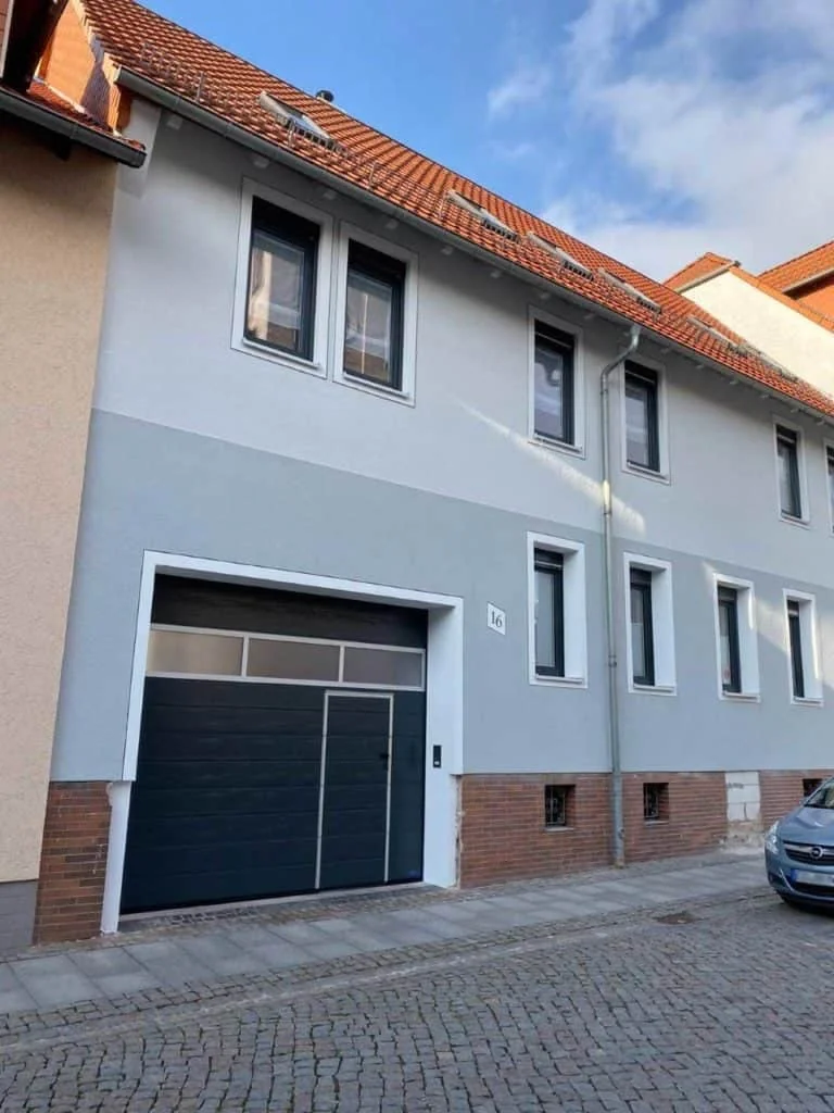 Haus mit grau abgesetztem Erdgeschoss, dunklem Garagentor und weißen Fensterlaibungen an einer gepflasterten Straße