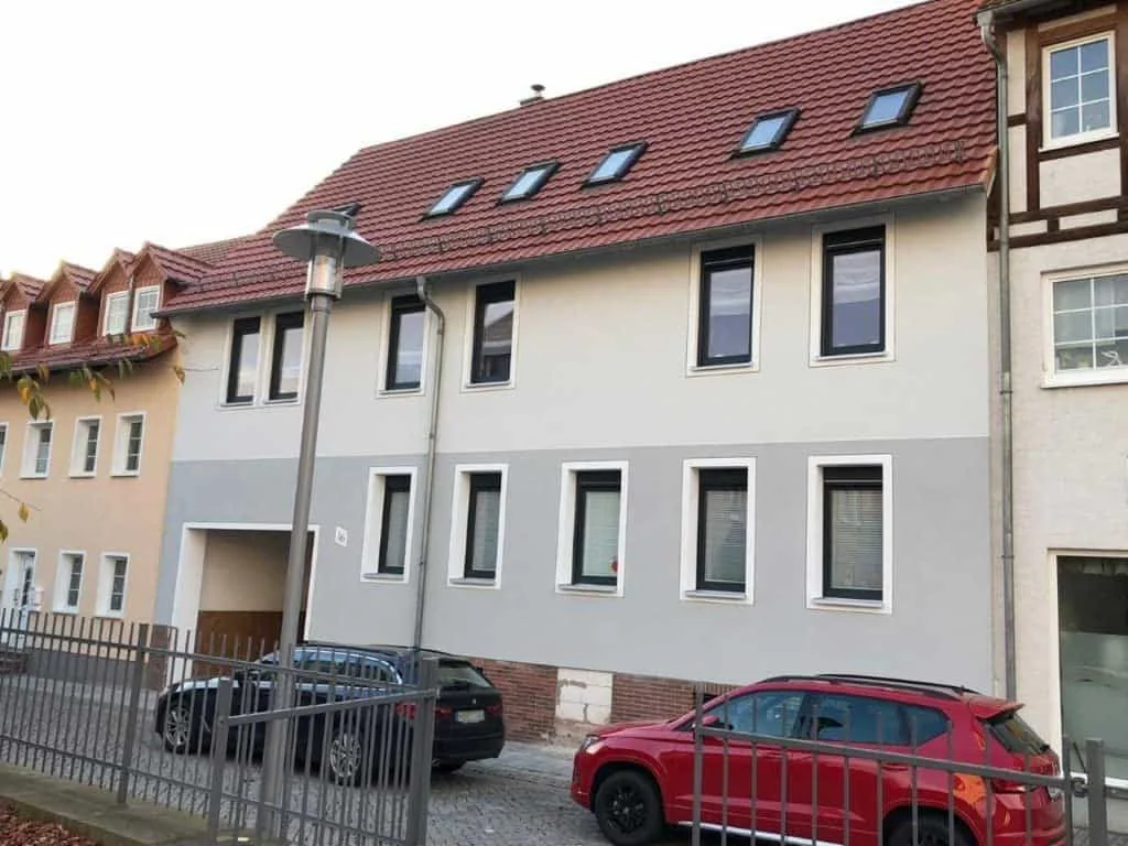 Haus mit grauer und weißer Fassade, Dachfenstern im roten Ziegeldach und Einfahrt, davor geparkte Autos hinter einem Zaun