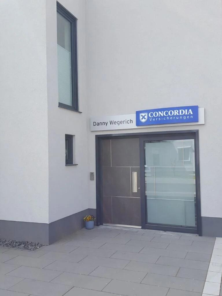 Eingang an einer hellen Hausecke mit dunkelbrauner Tür und breitem Glasseitenteil in grauem Rahmen